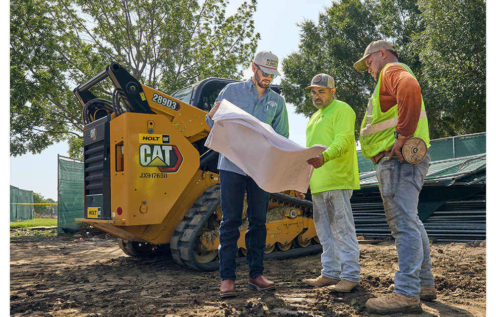 Three men, Nathan Menchaca and two workers, review blueprints at a job site beside a yellow CAT skid steer loader, with safety gear and work boots visible on a dirt-covered ground.